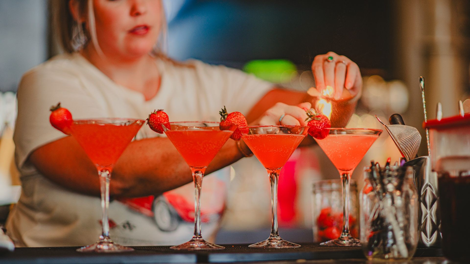 Bartender preparing strawberry cocktails with flaming garnish on a bar counter.