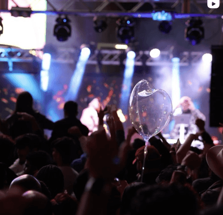 Crowd enjoying a concert with a heart-shaped balloon, stage lights illuminating the scene.