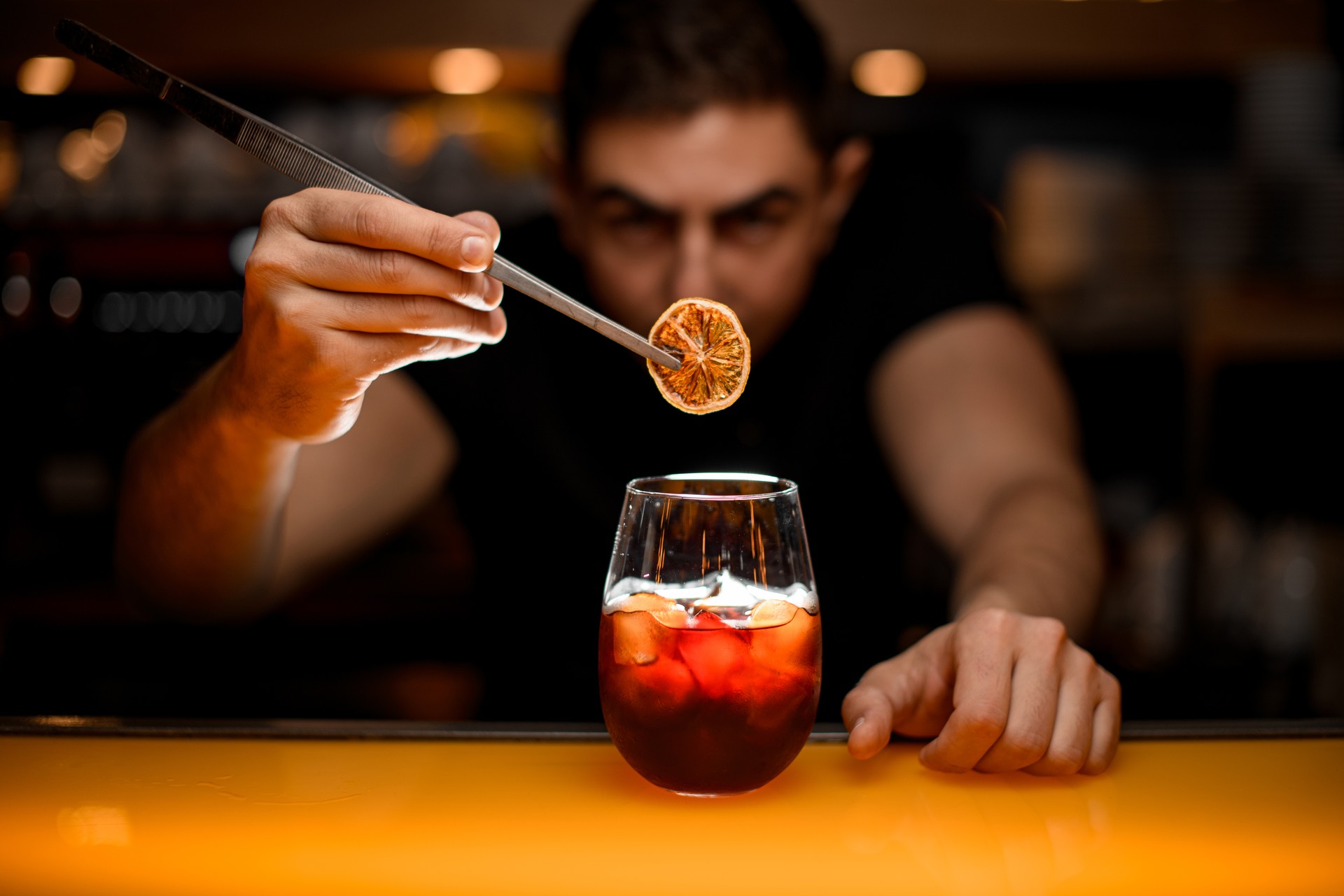 close-up of glass with cold drink and male hand accurate holding dry slice of orange with tweezers over it